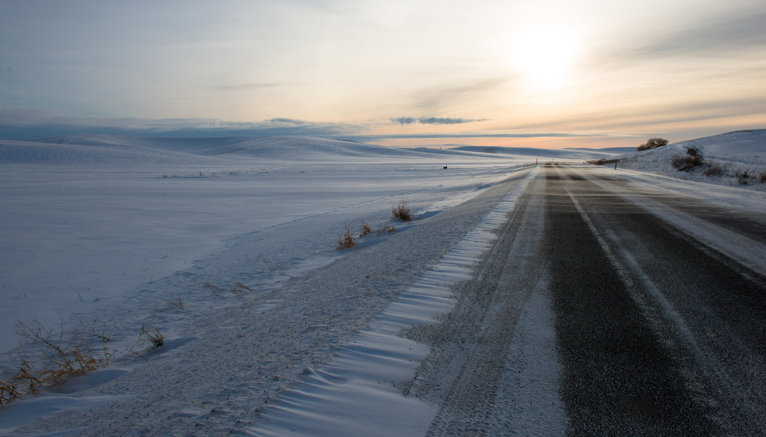 snowy landscape with road leading towards glimmer of sun