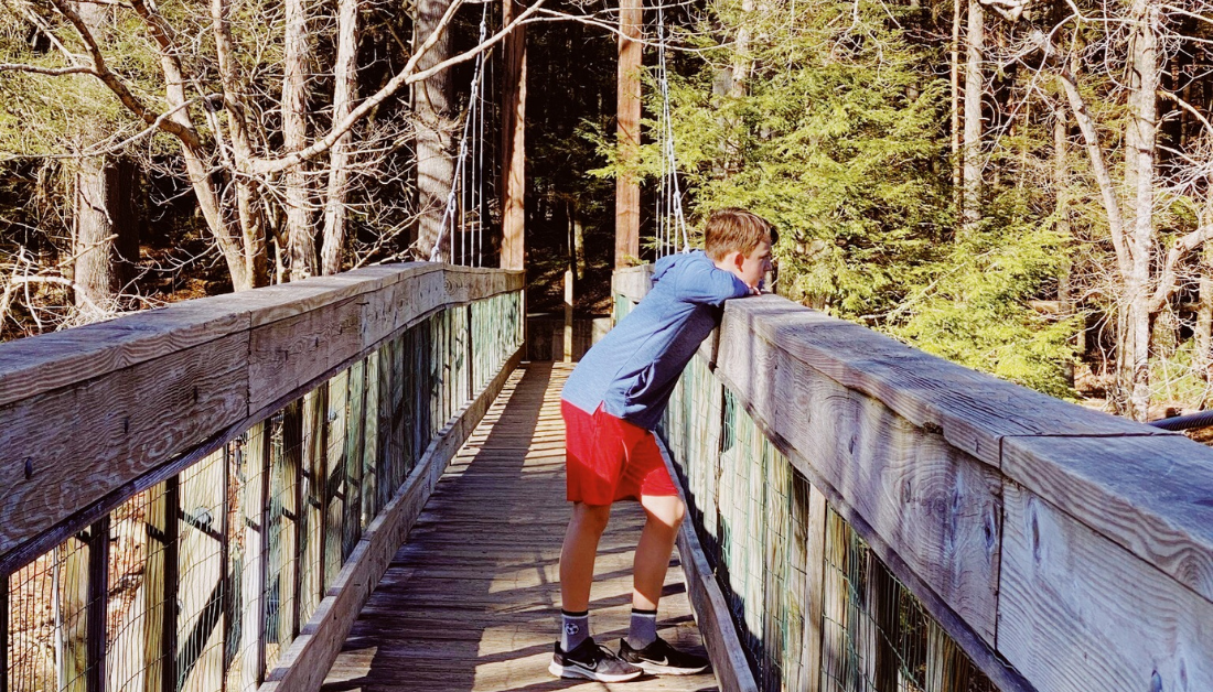 picture of boy leaning on the railing of a wooden bridge looking off camera with trees behind him