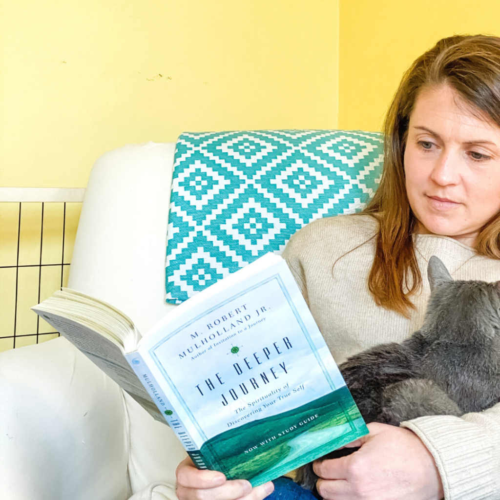 Amy Julia sitting in a white chair reading a book and holding a gray kitty