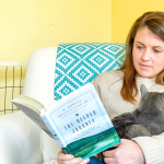 Amy Julia sitting in a white chair reading a book and holding a gray kitty