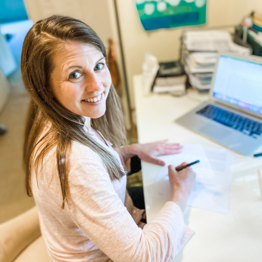 Amy Julia looking over her shoulder and smiling at the camera with a pen poised in her hand to sign a book contract