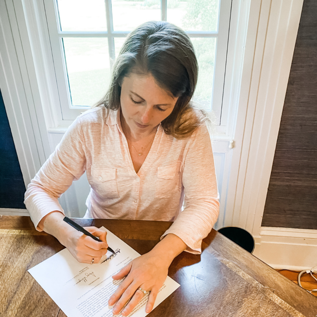 introduce myself picture of Amy Julia siting at a wooden table looking down signing a piece of paper with a window behind her