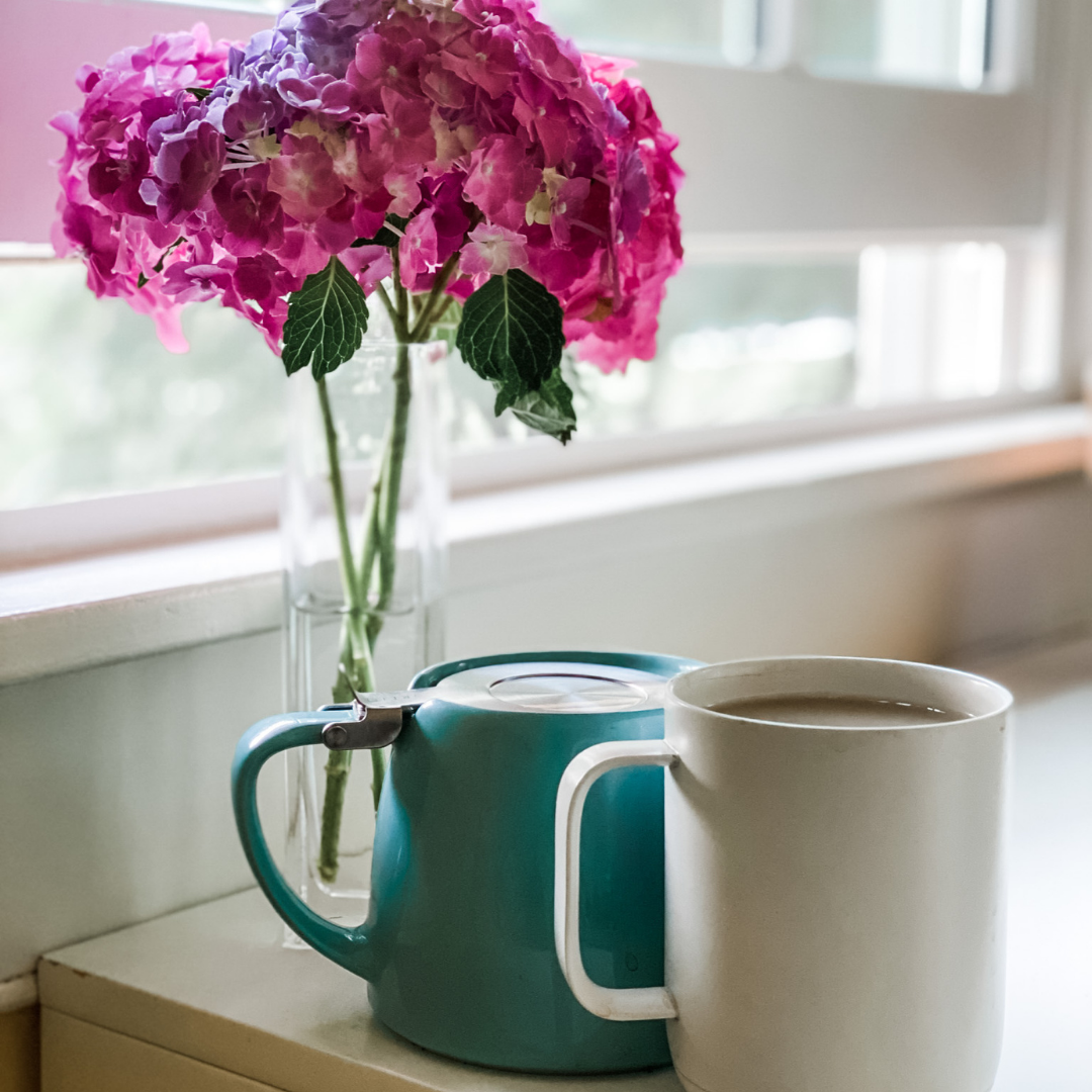 picture of pink flowers in a vase on a counter next to an open window and two mugs sitting next to the vase; one mug is greenish-blue and upside down and one mug is white and filled to the brim with tea; a peaceful scene for talking about emotional disability