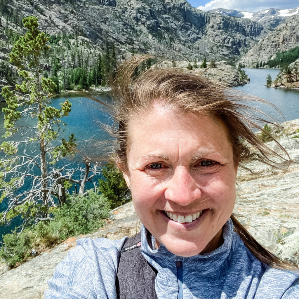 picture of Amy Julia smiling with welcome at the camera with a lake and mountains behind her