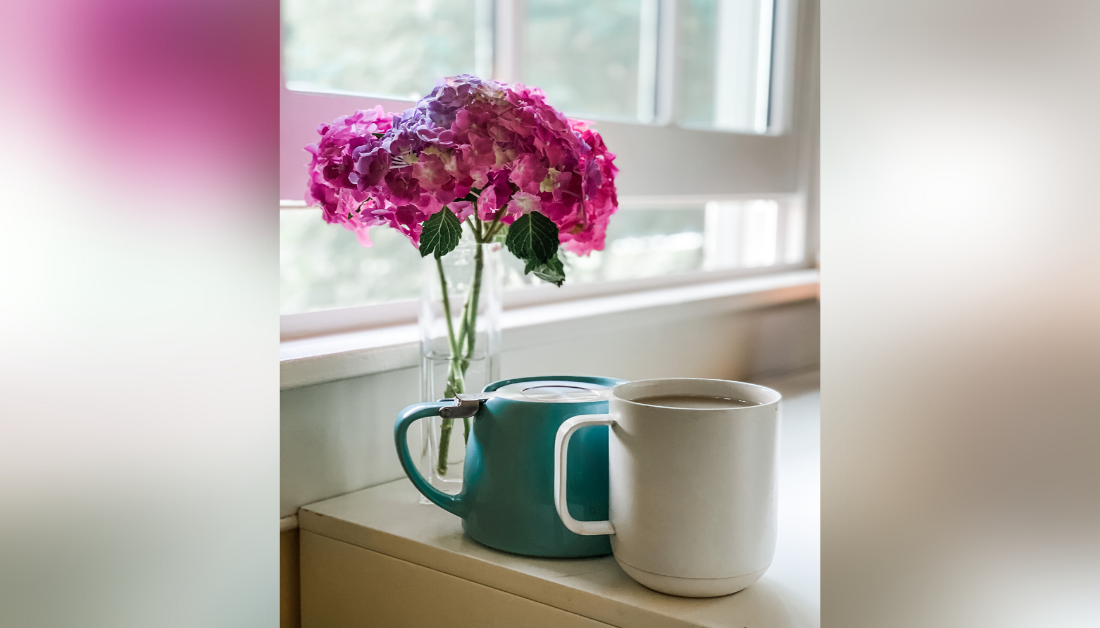 picture of pink flowers in a vase on a counter next to an open window and two mugs sitting next to the vase; one mug is greenish-blue and upside down and one mug is white and filled to the brim with tea