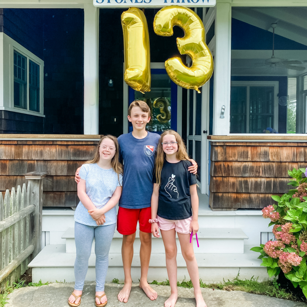 William standing in-between Penny and Marilee with his arms around them. All three are looking at the camera and smiling. There is a gold 13 balloon hanging from the porch in the background