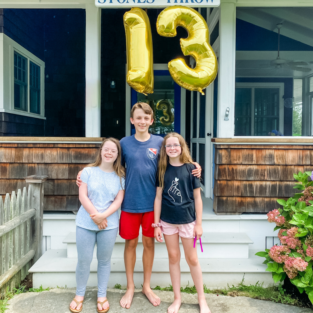 William standing in-between Penny and Marilee with his arms around them. All three are looking at the camera and smiling. There is a gold 13 balloon hanging from the porch in the background