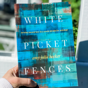 Amy Julia's hand holding the book White Picket Fences in front of a white picket fence in celebration of the book birthday for White Picket Fences