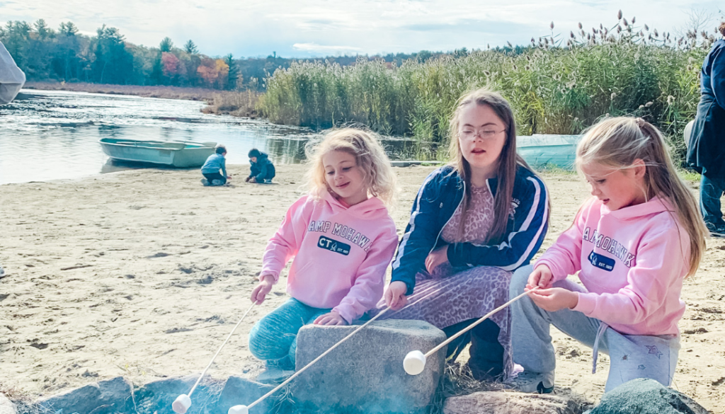 Three girls kneeling beside a campfire. They are roasting marshmallows. There is a lake, boat, and autumn forest in the background.