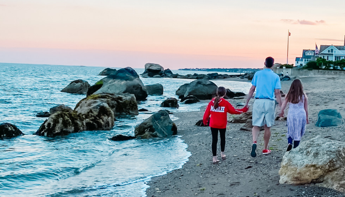 a grown up and two children walking away from the camera along the beach with water rippling around rocks to their left