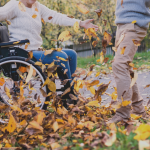 A couple on a walk through autumn leaves. One person is in a wheelchair. They are both throwing leaves.