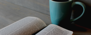 A Bible sits on a wooden table next to a blue mug filled with coffee.