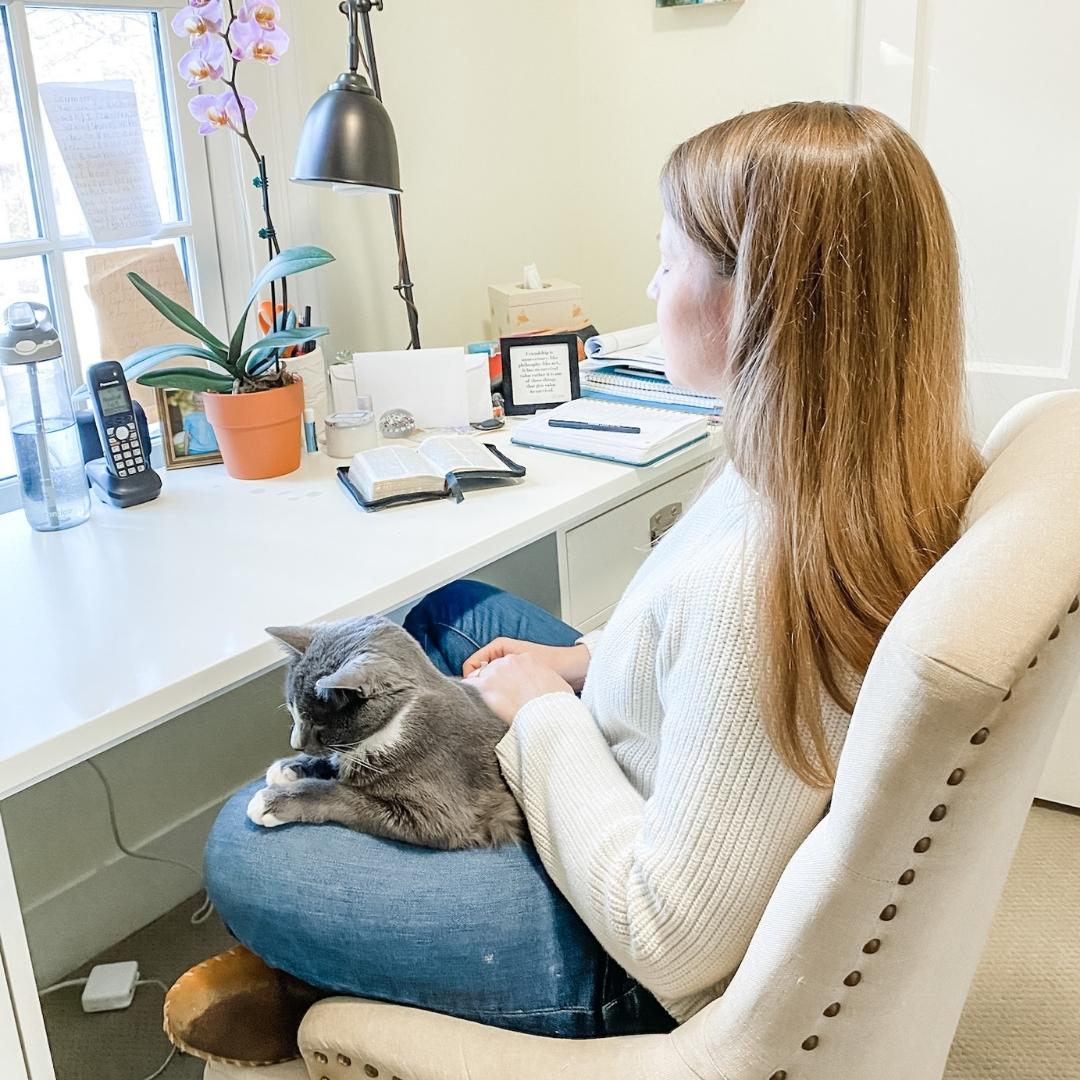 Amy Julia sits in a chair at her desk. Her eyes are closed and she is holding a gray cat.