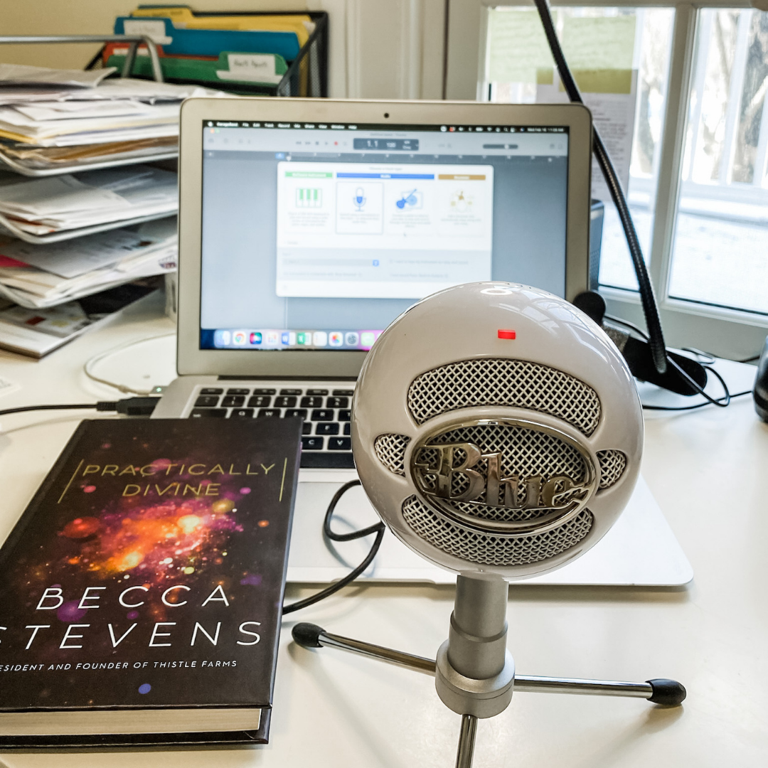 Amy Julia's desk with a podcast mic, computer, and the book Practically Divine