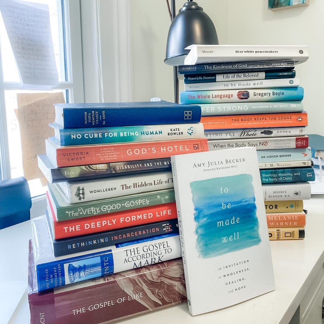 stack of books on Amy Julia's desk with the book To Be Made Well leaning against the stack