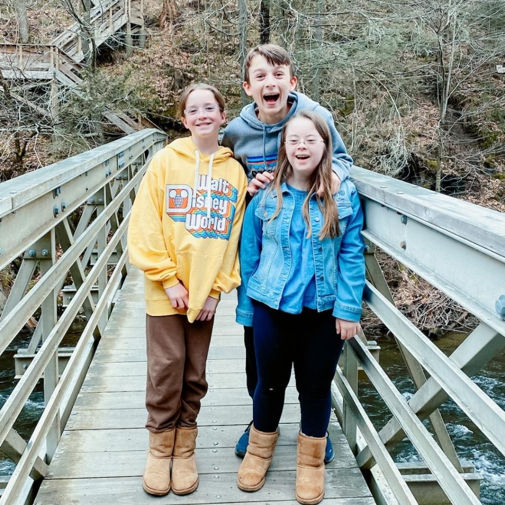 Marilee, William, and Penny smile happily at the camera while they stand on a wood bridge with a forest hill in the background
