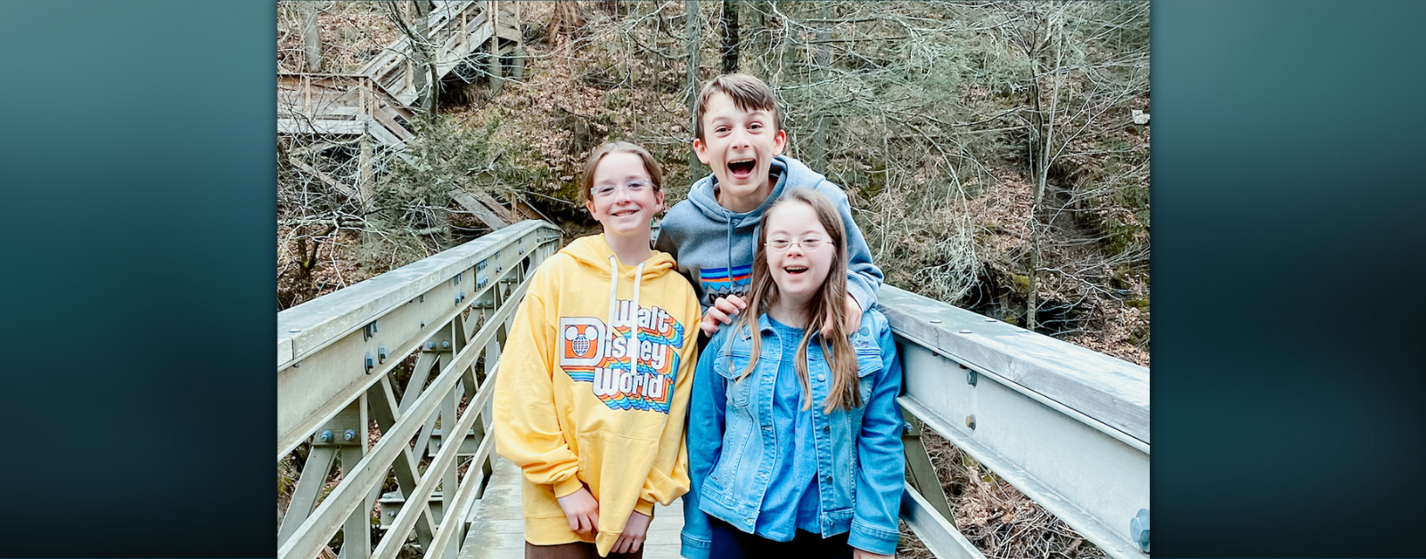 Marilee, William, and Penny smile happily at the camera while they stand on a wood bridge with a forest hill in the background