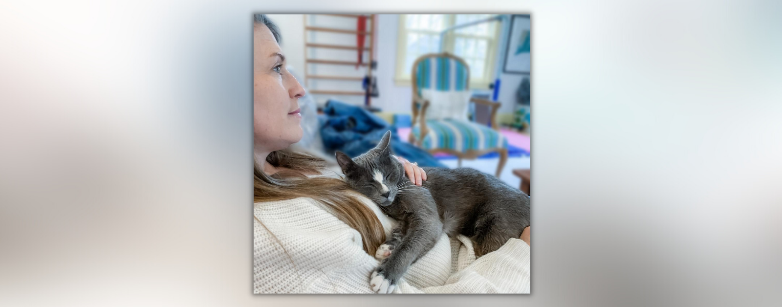 Amy Julis looks across the room while she sits in a chair and holds her gray kitty on her chest