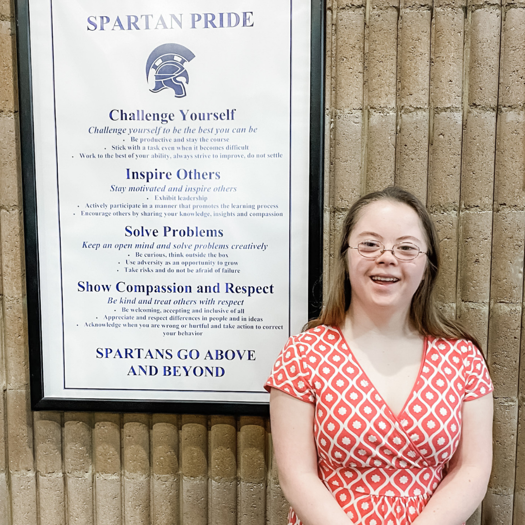 Penny smiles at the camera and stands in front of a wall with a framed list of school values