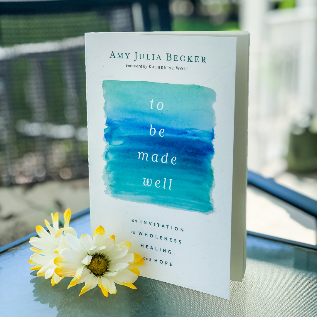 a copy of To Be Made Well stands on a glass table. White and yellow-tipped flowers are on the table in front of the book