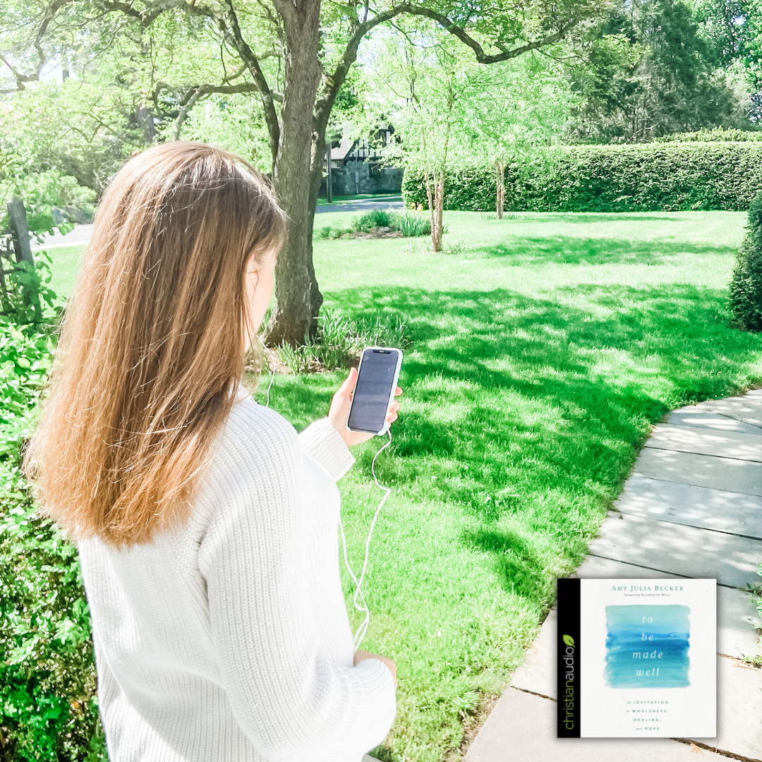 Amy Julia stands outside with her back to the camera. A green lawn and wooden walkway stretch out in front of her. She is holding a phone and has earphones in.