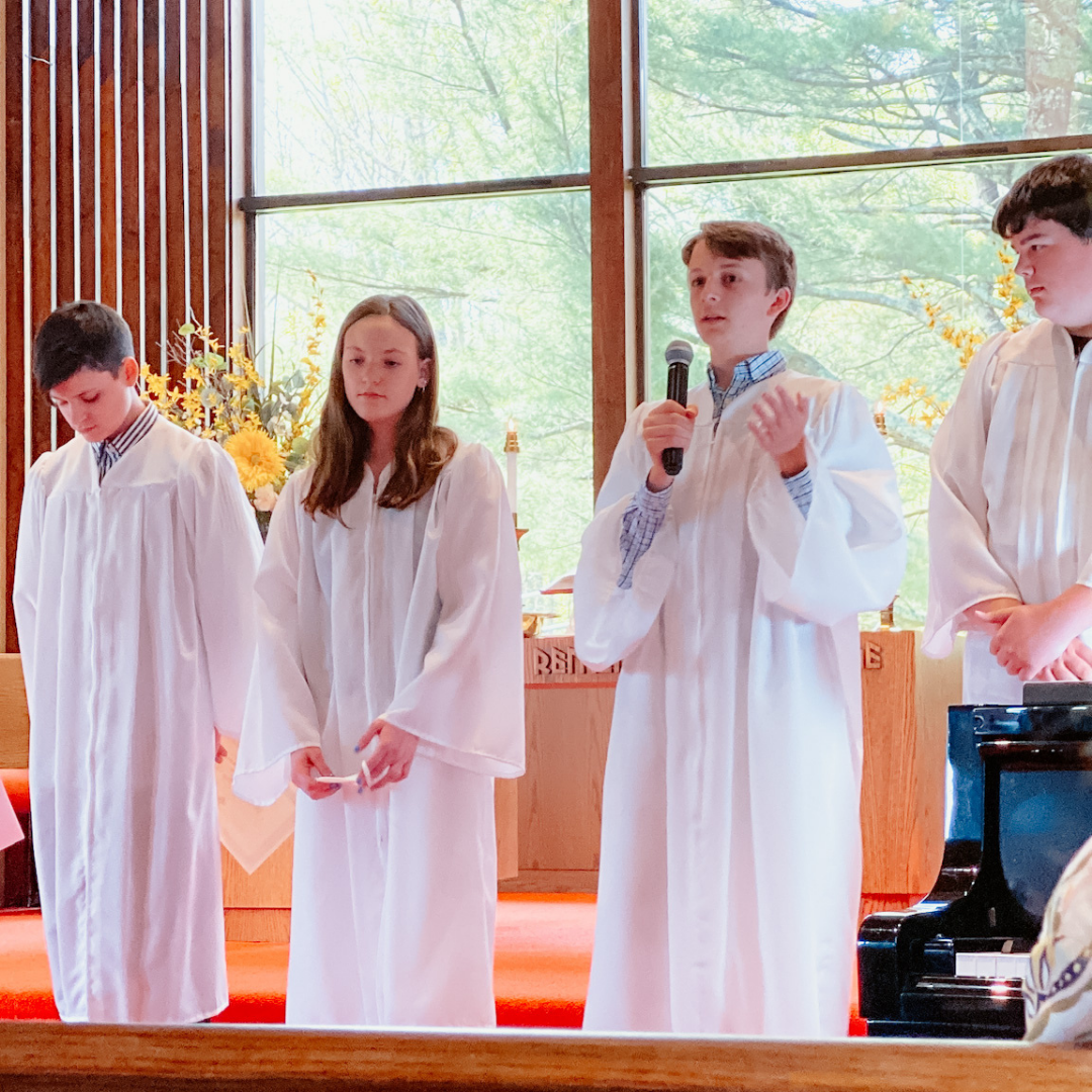 William stands at the front of church and speaks into a mic he holds in his hand. He, and the teens standing next to him, are wearing white robes.