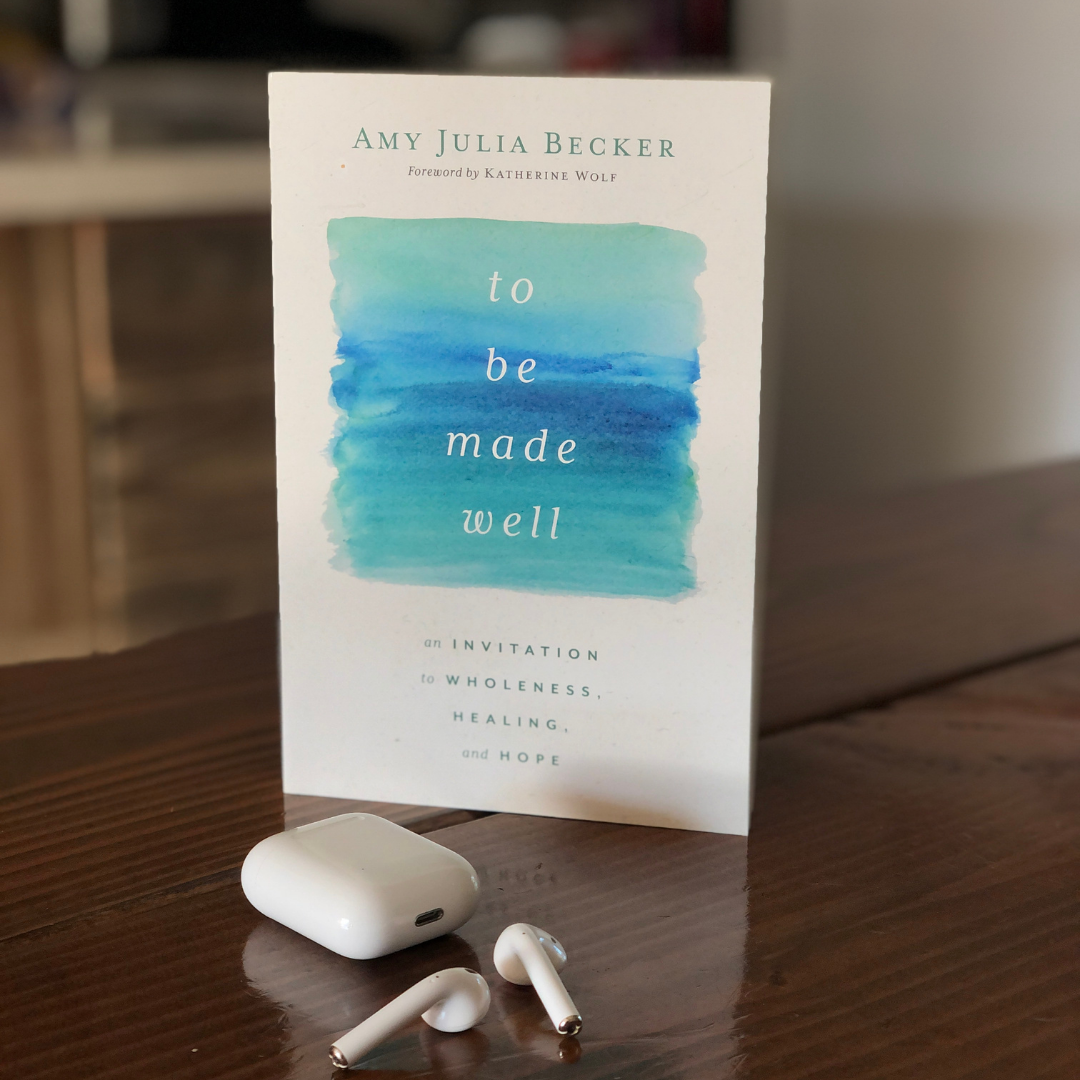 a copy of To Be Made Well stands on a wooden table with an airpod case and airpods on the table in front of the book