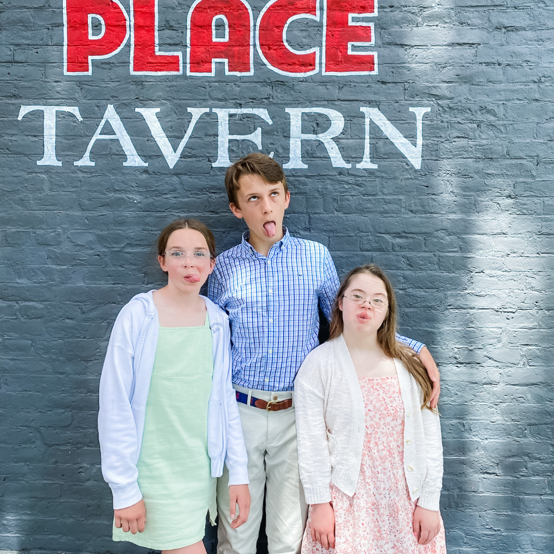 Marilee, William, and Penny make funny faces at the camera and stand in front of a gray stone wall. Williams arms are around his sisters' shoulders.