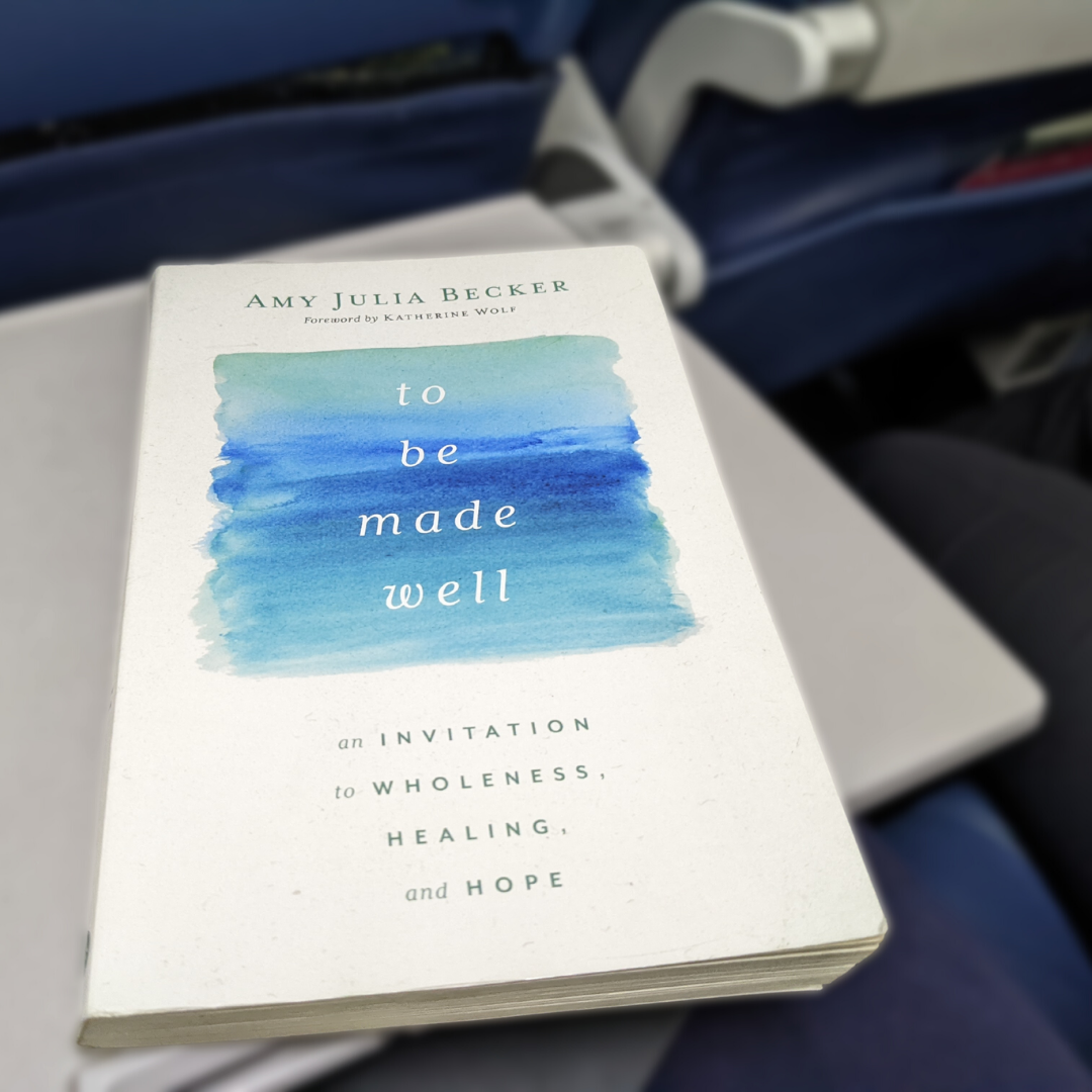 a copy of To Be Made Well rests on a seat tray on an airplane