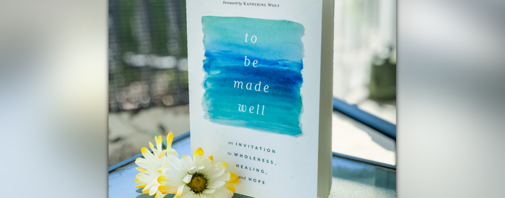 a copy of To Be Made Well stands on a glass table. White and yellow-tipped flowers are on the table in front of the book