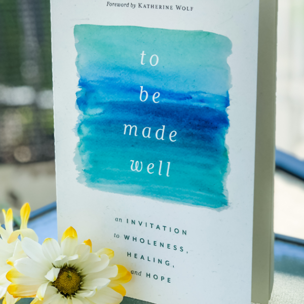 a copy of To Be Made Well stands on a glass table. White and yellow-tipped flowers are on the table in front of the book