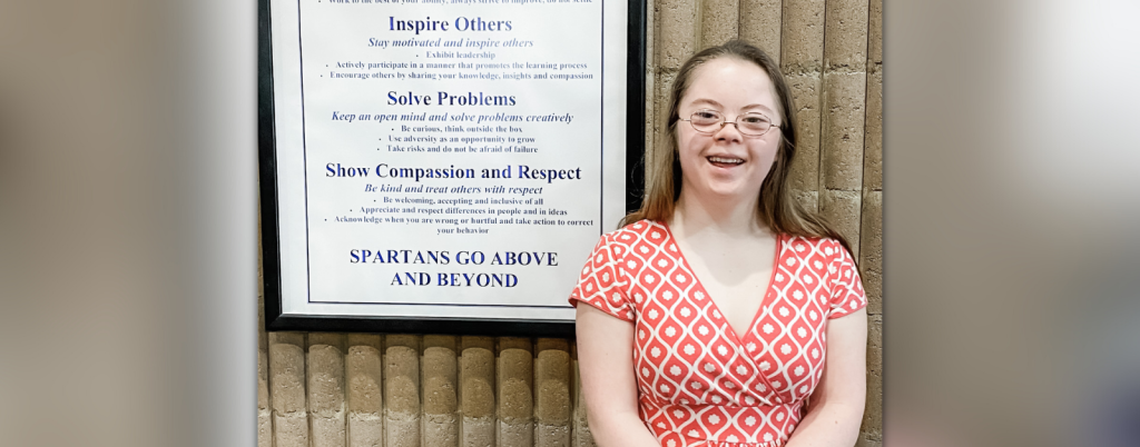 Penny smiles at the camera and stands in front of a wall with a framed list of school values