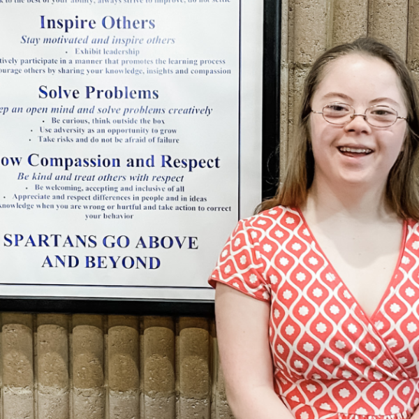 Penny smiles at the camera and stands in front of a wall with a framed list of school values