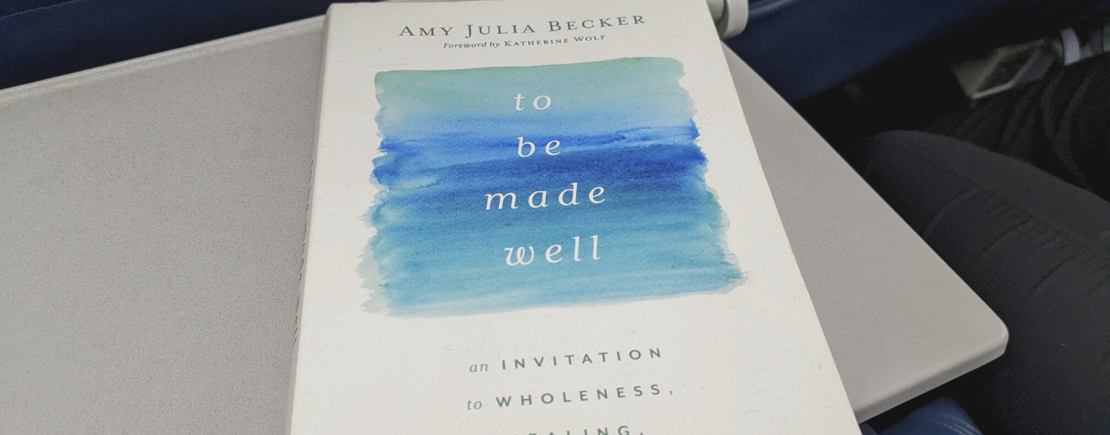 a copy of To Be Made Well rests on a seat tray on an airplane