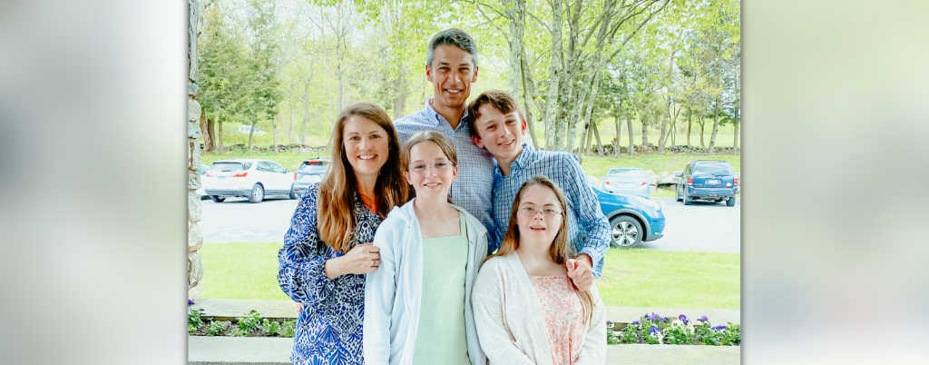 Becker family stands outside in front of a parking lot and low stone wall. They smile at the camera