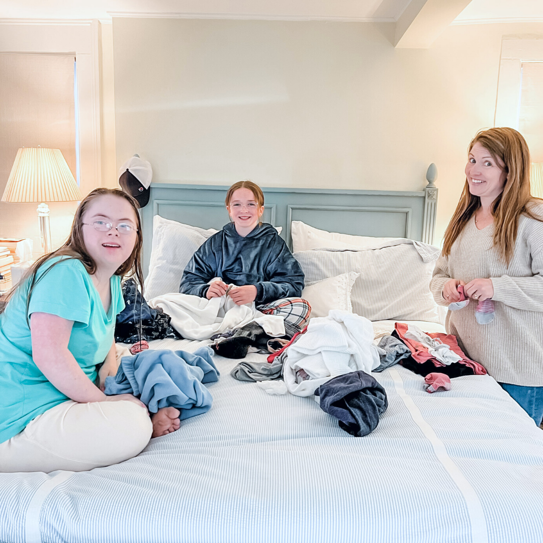 Penny and Marilee sit on a white bed and Amy Julia stands next to it. They are folding clothes and smiling at the camera.