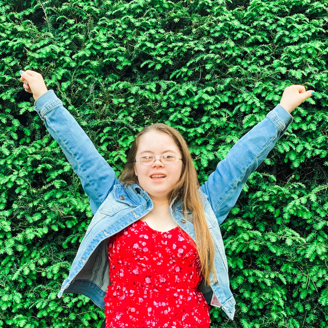 Penny raises her arms in a cheer in front of a hedge of greenery