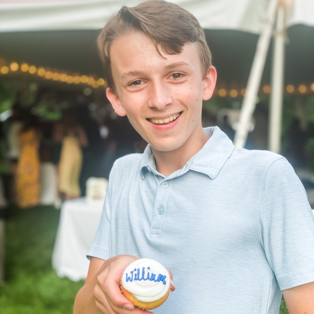 William smiles and stands in front of a tent and holds a cupcake with white frosting and "William" written in blue frosting