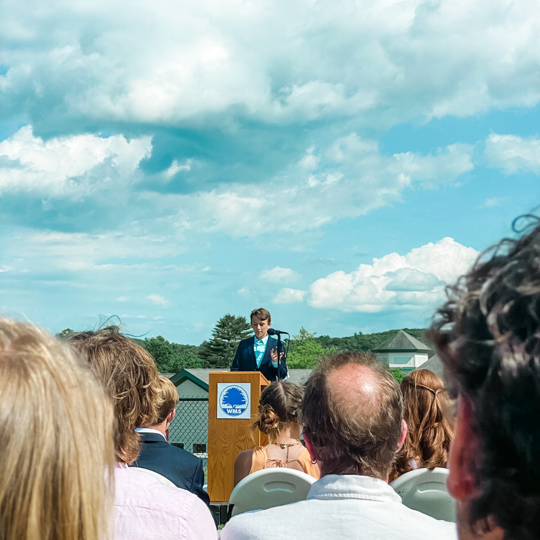 William stands at an outdoor podium and gives a speech with blue sky and clouds behind him