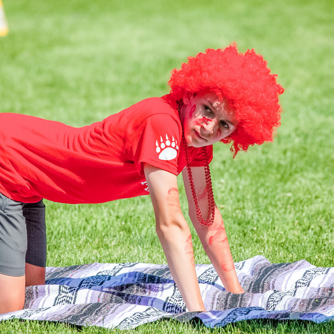 William kneels on a blanket in the grass. he is wearing a red t-shirt and wig.