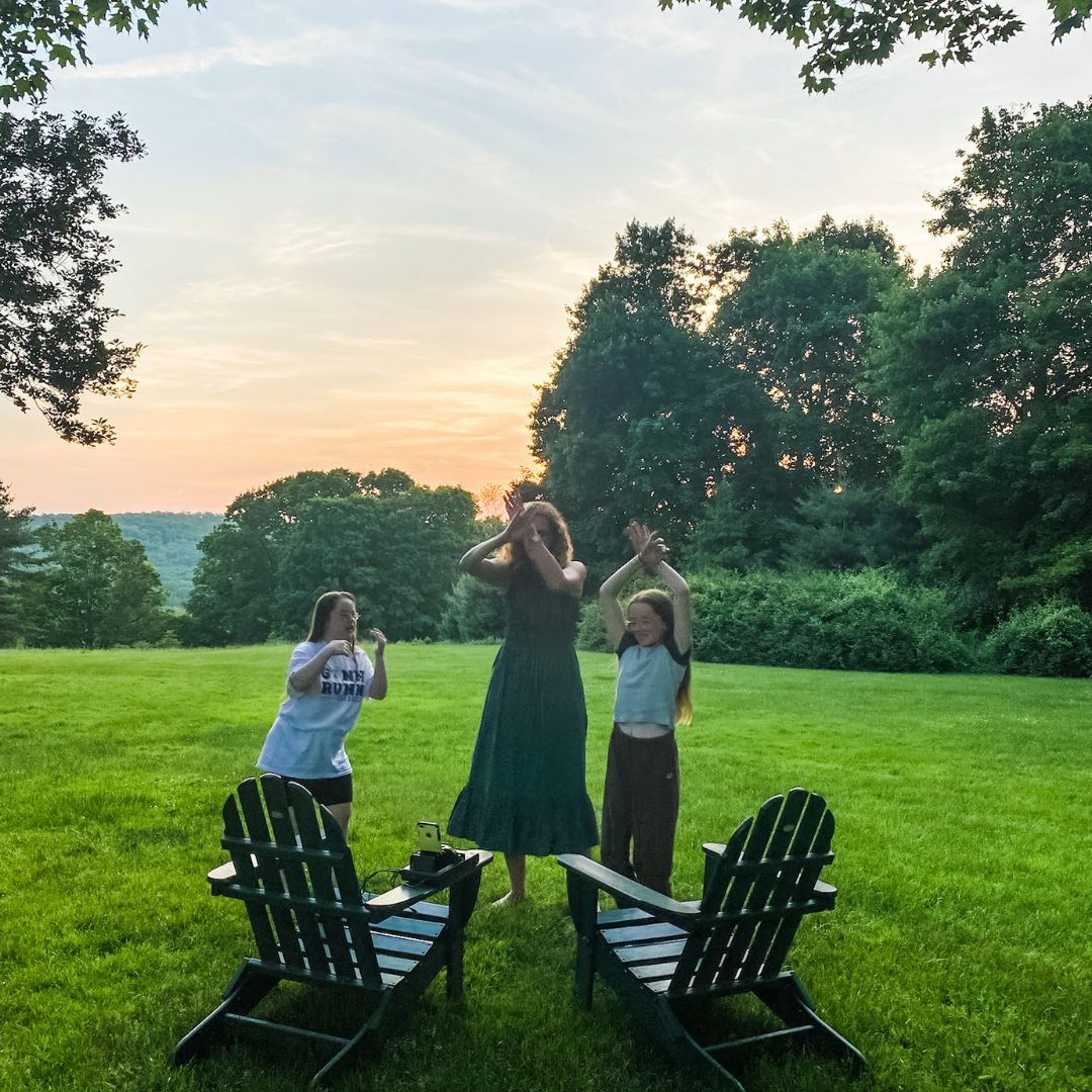 Penny, Elizabeth, and Marilee dance to a TikTok video in front of lawn chairs and with trees and a glowing sunset in the background