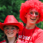 William stands behind Marilee with his arms around her. They are both wearing red t-shirts. Marilee wears a red hat, and William wears a red wig.
