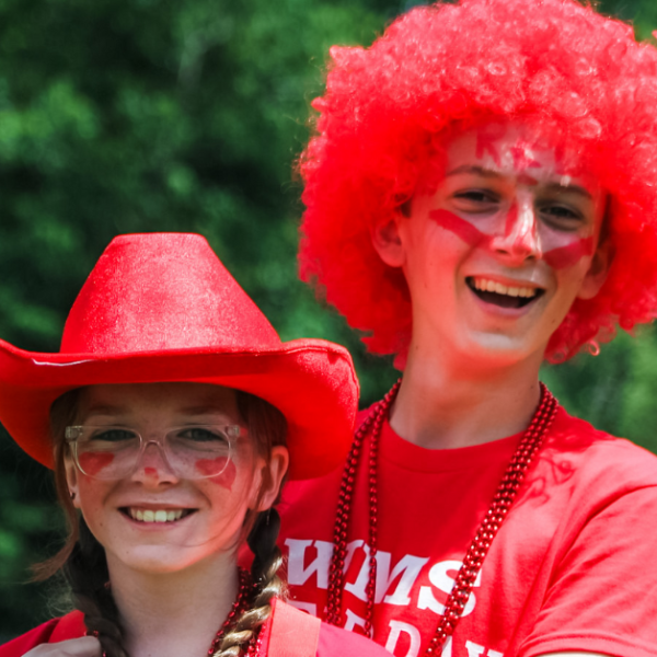 William stands behind Marilee with his arms around her. They are both wearing red t-shirts. Marilee wears a red hat, and William wears a red wig.