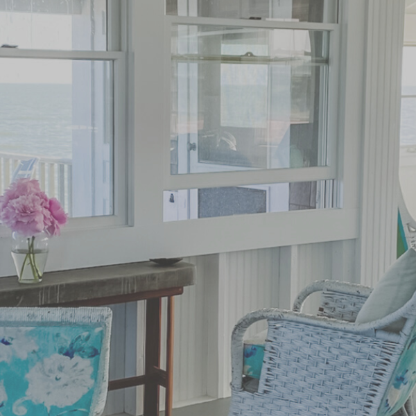 pink flowers on a table in a beach house in front of a window looking out at the ocean and surrounded by chairs