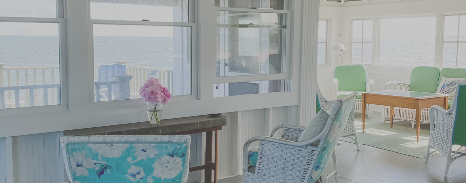 pink flowers on a table in a beach house in front of a window looking out at the ocean and surrounded by chairs