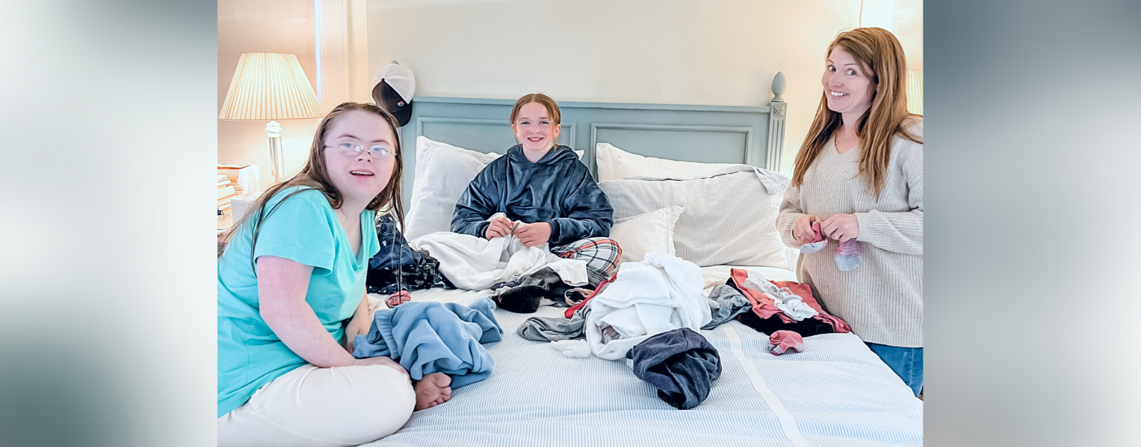 Penny and Marilee sit on a white bed and Amy Julia stands next to it. They are folding clothes and smiling at the camera.