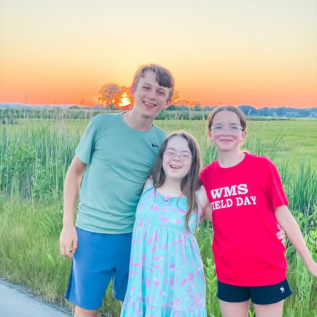 William, Penny, and Marilee smile at the camera with their arms around each other. They stand in front of tall green grass and a sunset in the background.