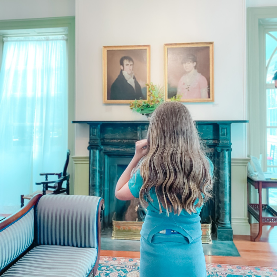 Marilee stands with her back to the camera in an old fashioned living room and looks at portraits on a wall above a fireplace