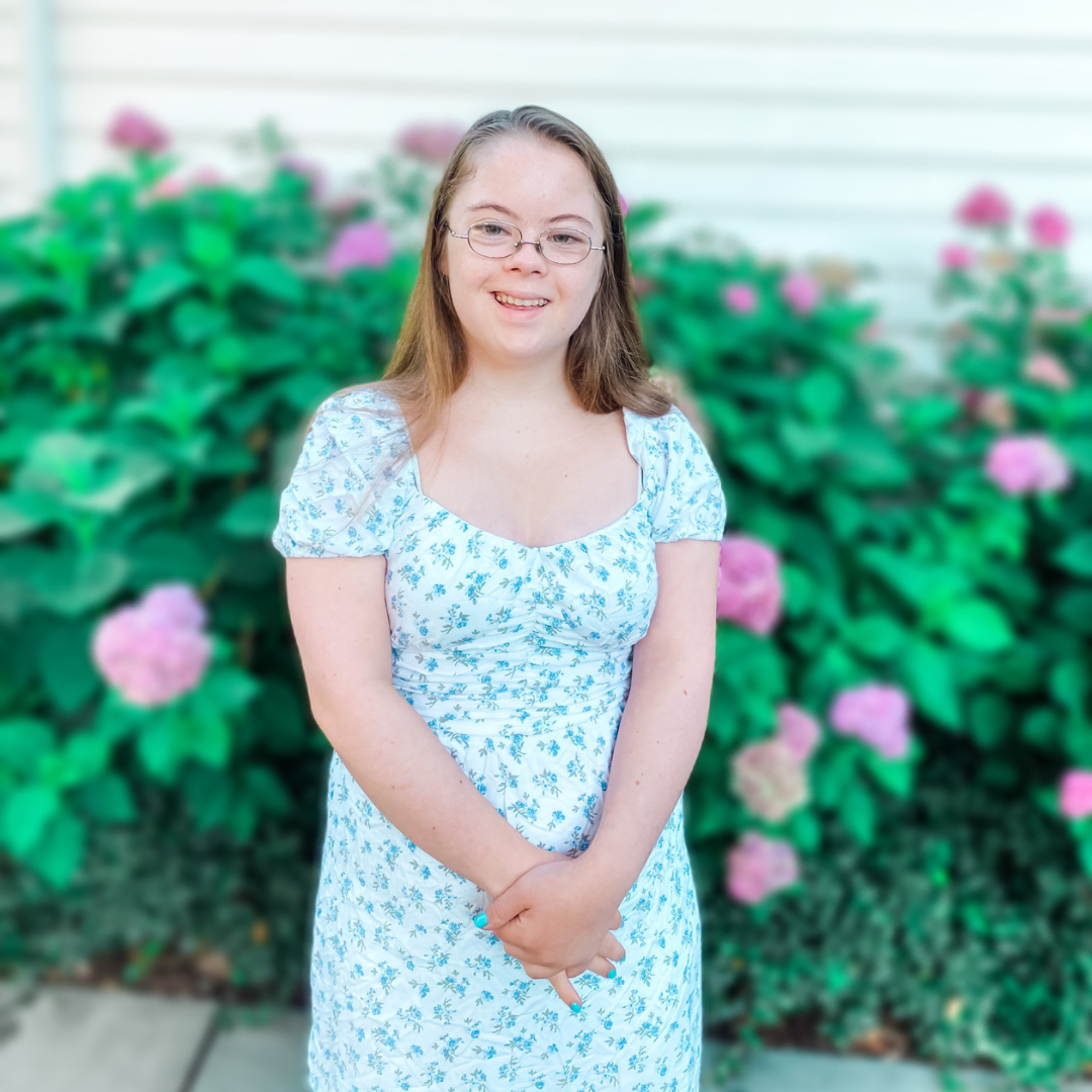 Penny is wearing a dress and stands in front of flower bushes as she smiles at the camera with her hands clasped in front of her.