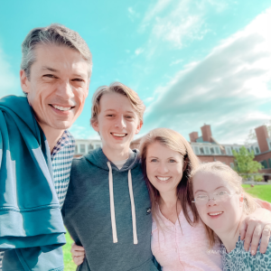 Peter, William, Amy Julia, and Penny smile for a selfie with their arms around each other and stand in front of a brick building with a brilliant blue sky behind them